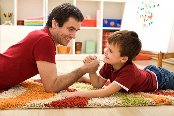 A father lies on a rug face to face with his young son, both laughing, with a stack of toys between them.